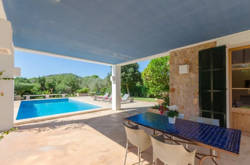 Una zona de comedor exterior cubierta da a una piscina y un jardín verde.