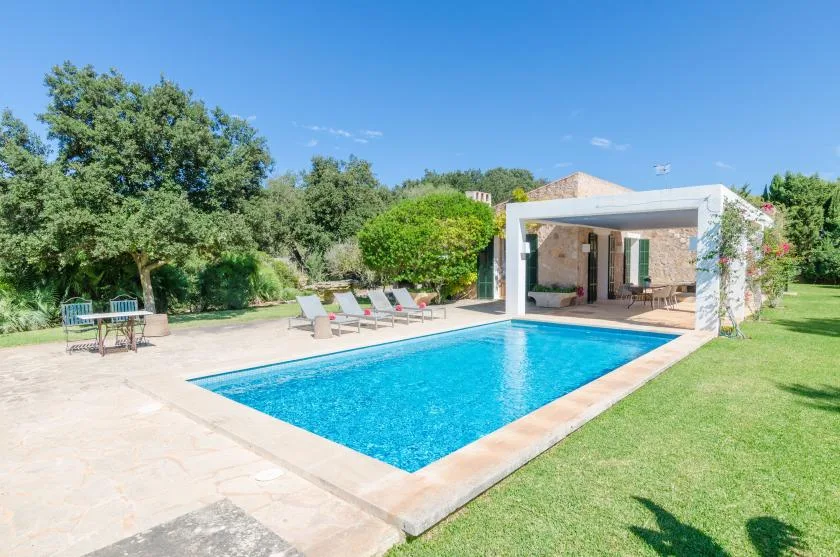 Una piscina al aire libre junto a un patio cubierto, con césped y árboles.