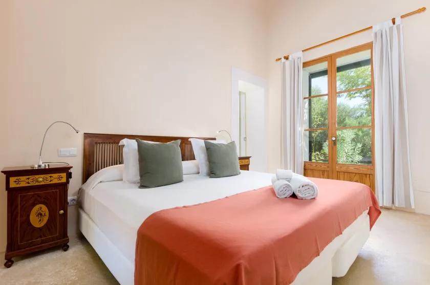 Un dormitorio con una cama bien tendida y ventanas altas enmarcadas por cortinas blancas.