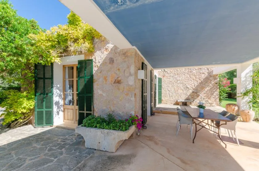 Un patio de piedra a la sombra con una mesa de comedor y contraventanas verdes.