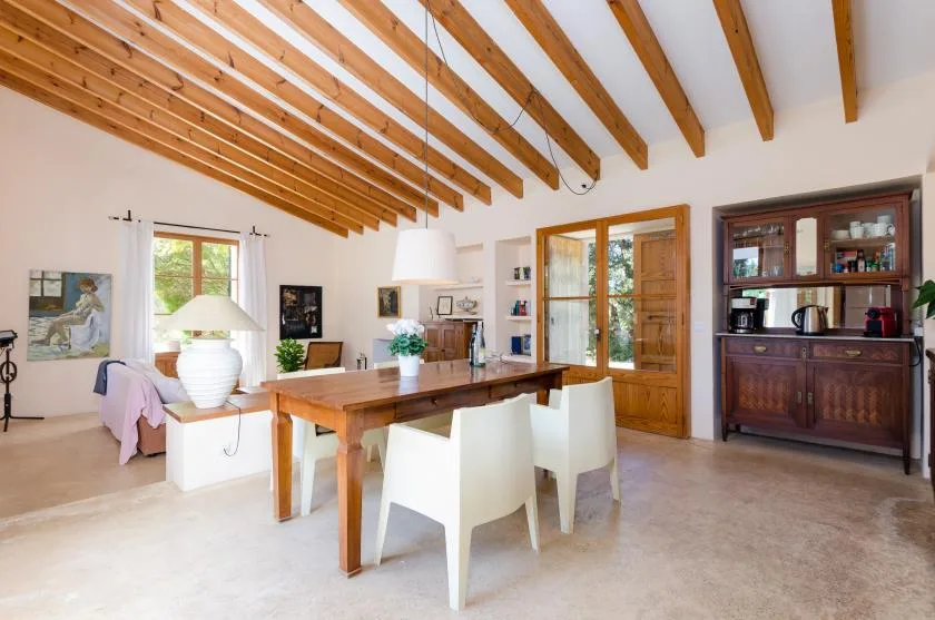 Comedor con una mesa de madera y asientos blancos bajo un techo con vigas vistas.
