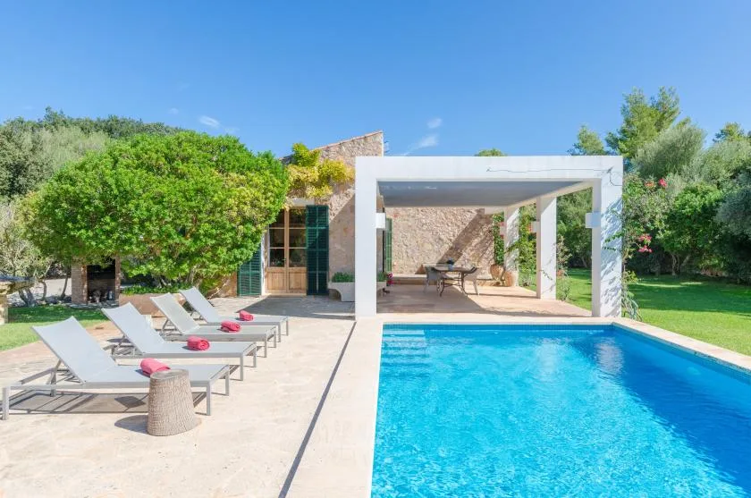 Una piscina junto a un patio con tumbonas y una zona de estar a la sombra.