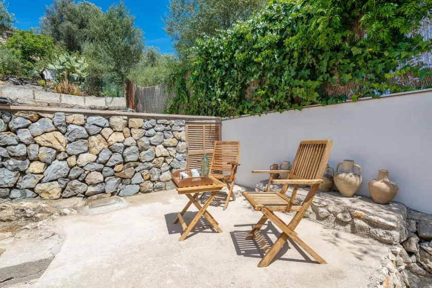 An outdoor seating area with wooden chairs beside a stone wall and leafy greenery.