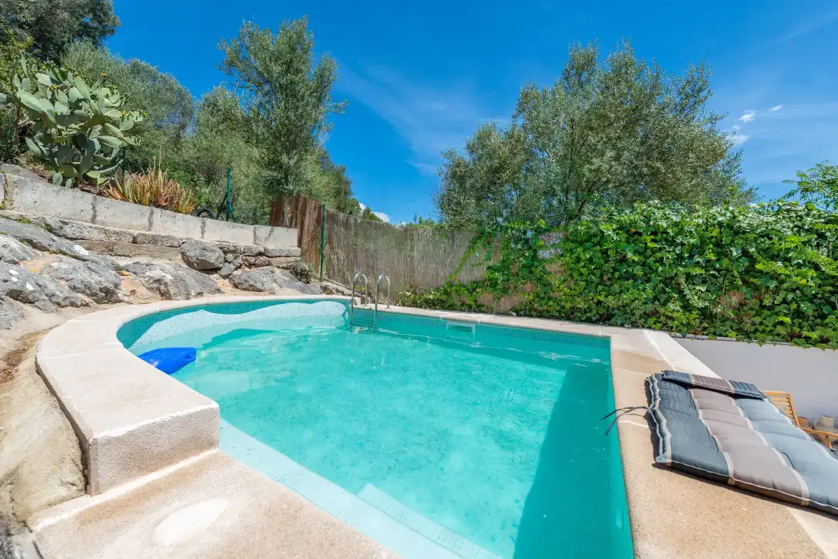 An outdoor swimming pool sits beside a stone retaining wall and leafy greenery.
