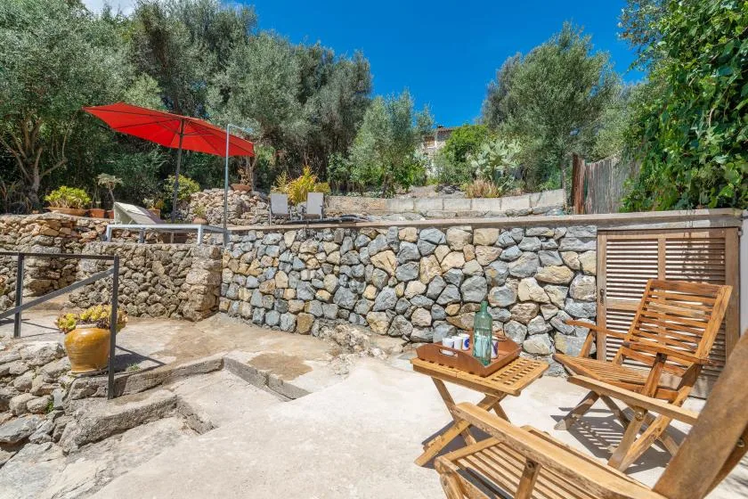 A sunny stone patio with wooden seating, a red umbrella, and surrounding trees.
