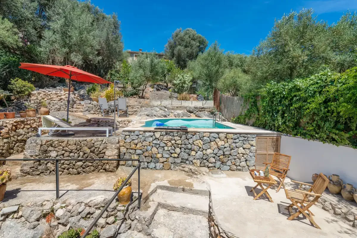 A stone terrace surrounds a swimming pool, with outdoor seating and a red umbrella.