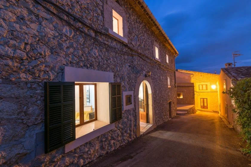 A stone building beside a narrow lane at dusk, with illuminated windows.