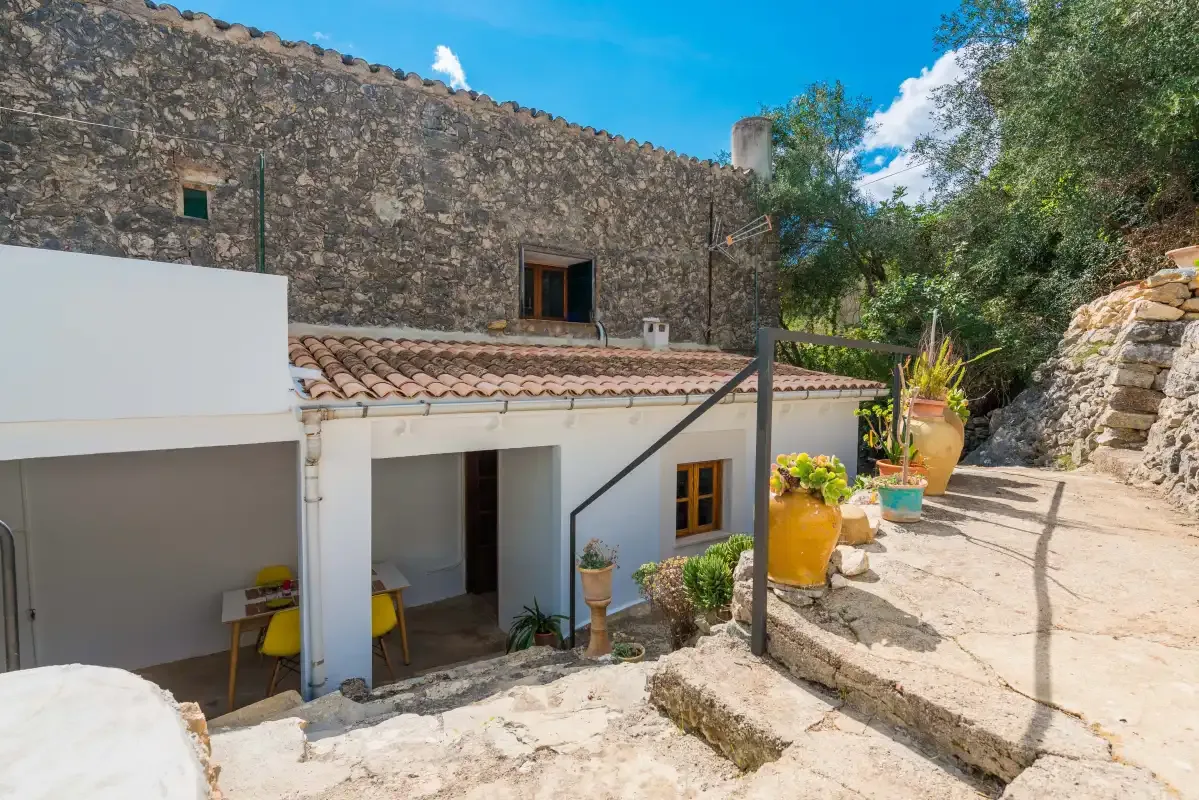A stone house opens onto a sunny outdoor terrace with potted plants.