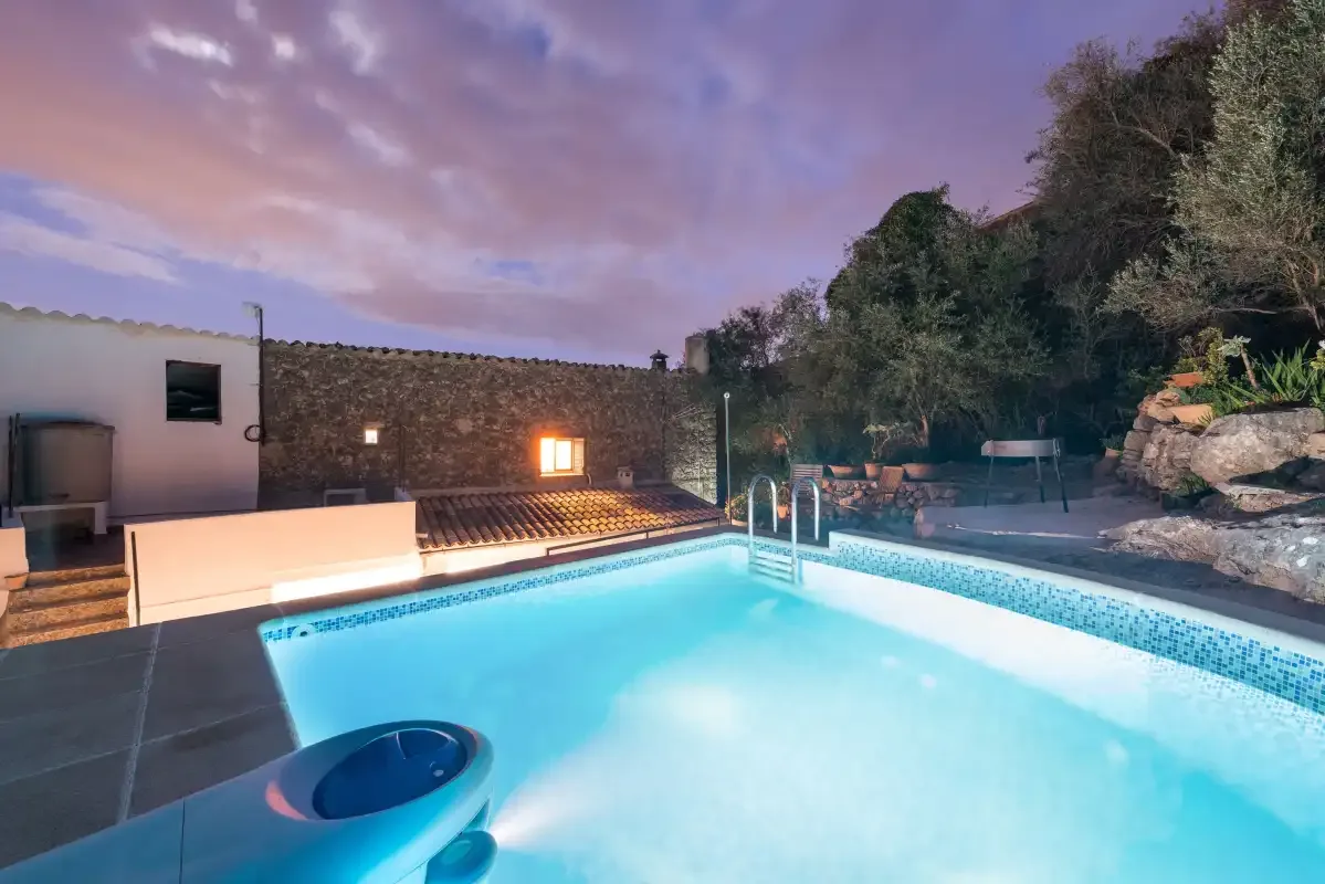 An outdoor swimming pool glows beside a stone wall at dusk.