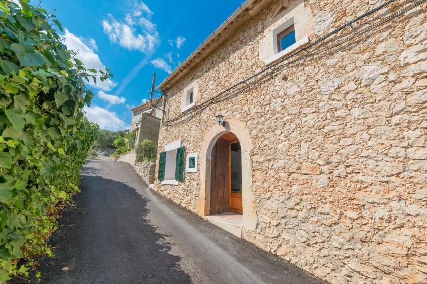 A narrow paved lane passes a stone building with an arched doorway and green shutters.