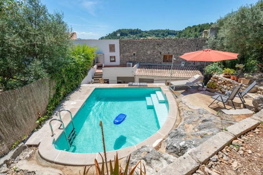 A turquoise swimming pool sits beside a stone patio with loungers and a red umbrella.
