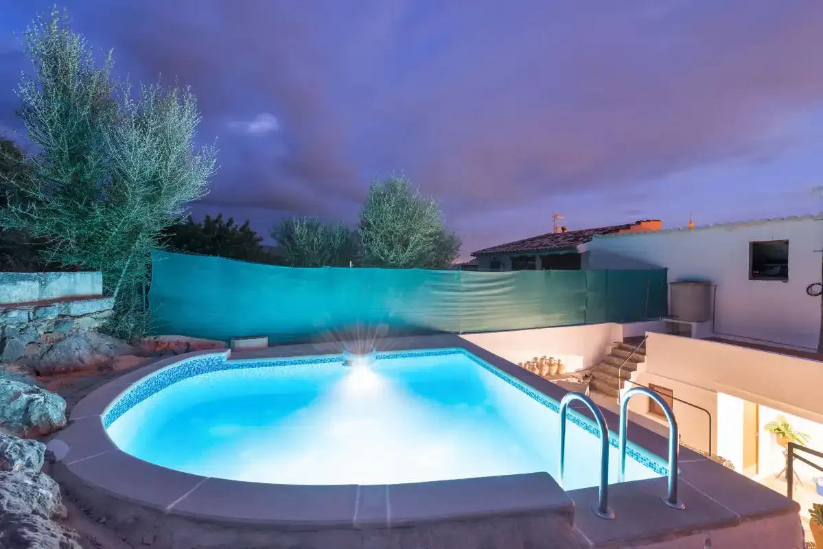 An illuminated outdoor pool sits beside a low house under a dusky sky.