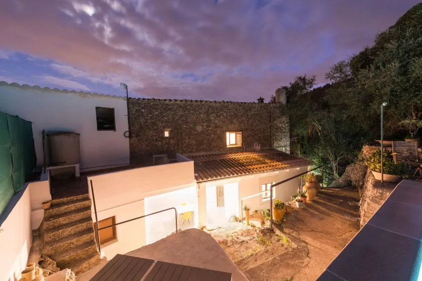 A courtyard at dusk with white buildings, stone walls, and surrounding trees.