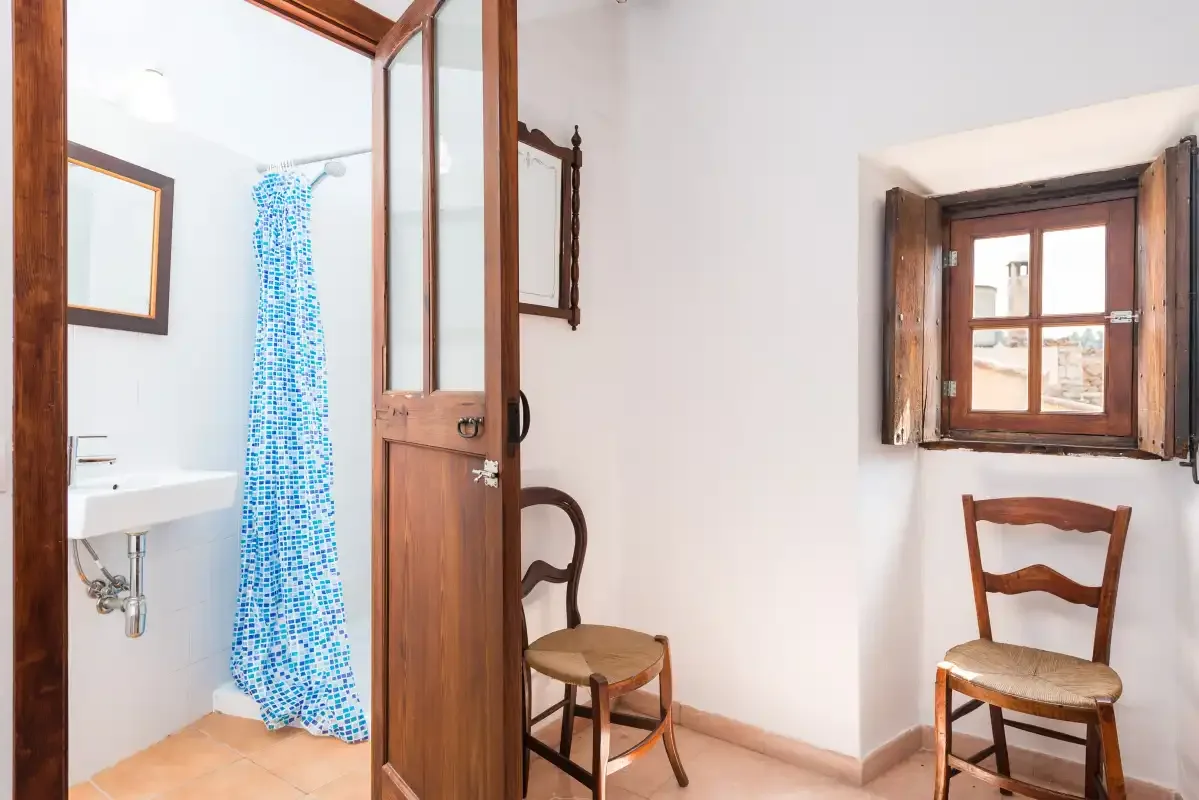 A bright room with a washbasin, patterned shower curtain, and wooden chairs.