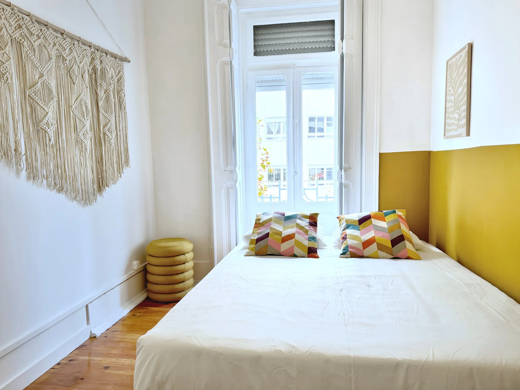 A bright bedroom with a bed, colorful cushions, and a tall window.