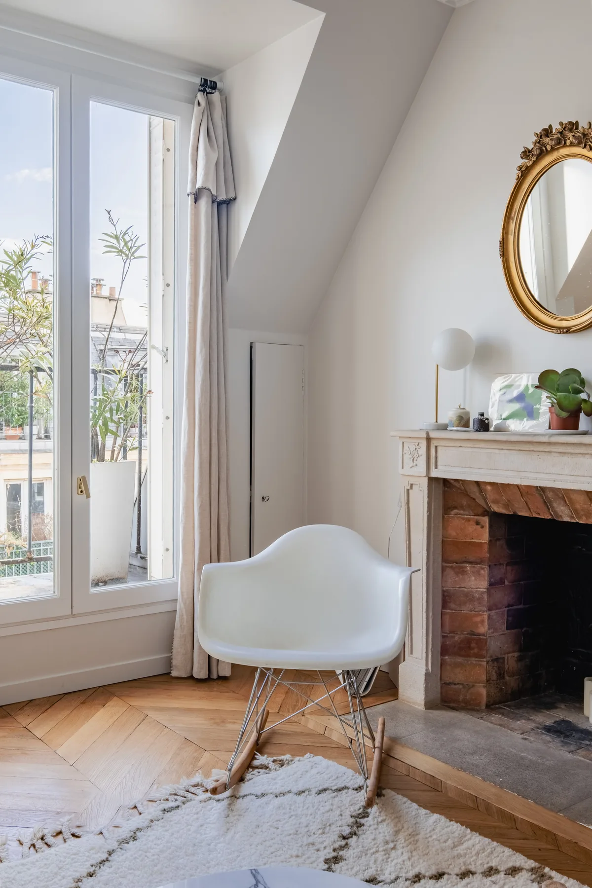 A bright sitting area with a white chair beside a brick fireplace and tall windows.