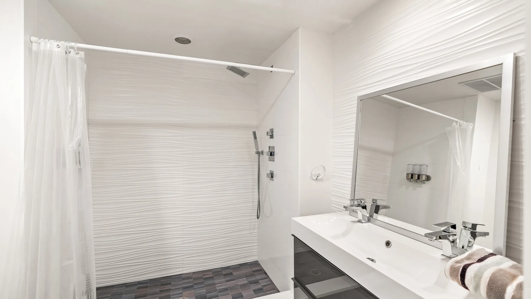 Un baño luminoso con ducha, cortina y lavabo bajo un espejo grande.