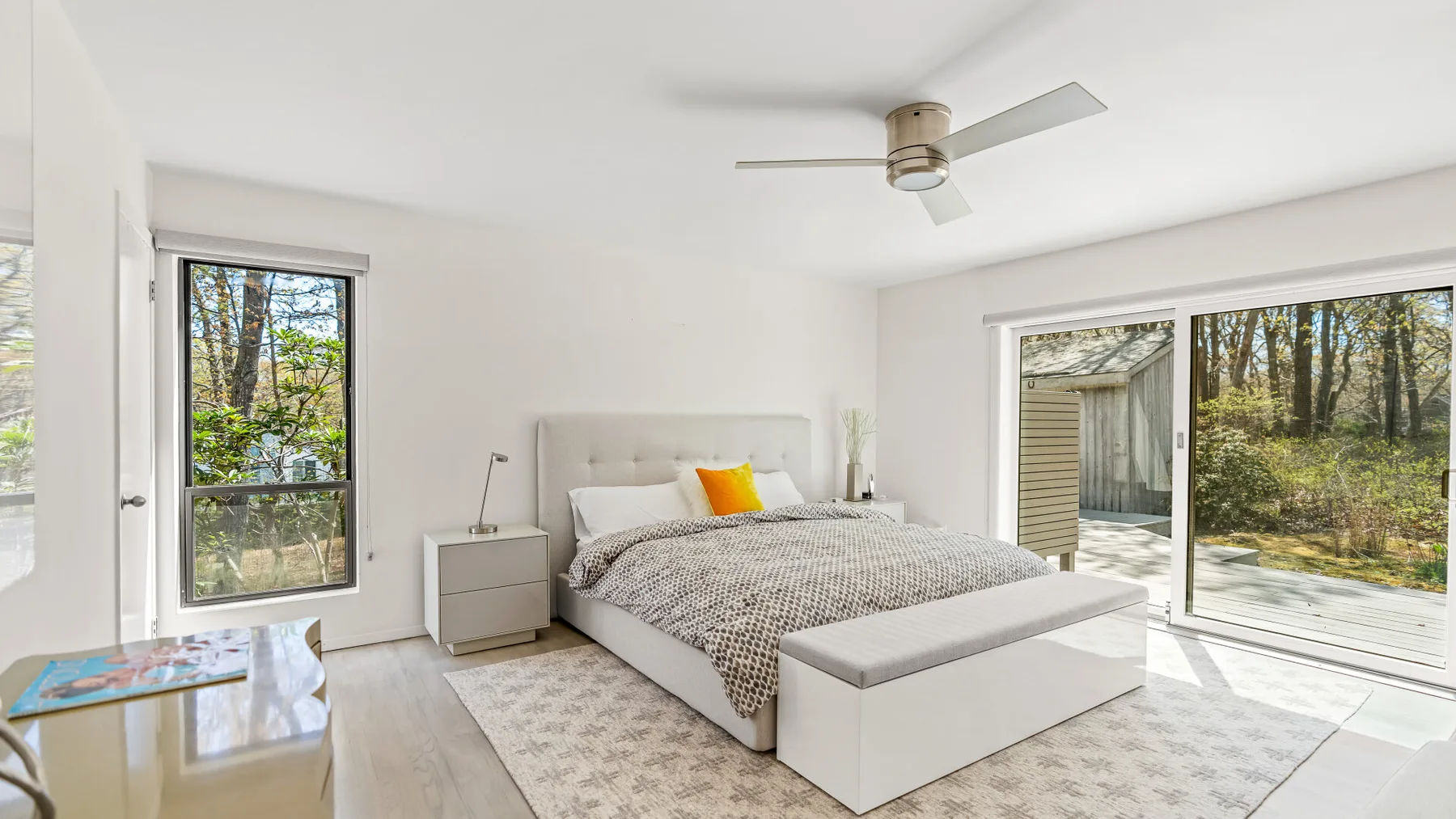 Un dormitorio luminoso con una cama, mesitas de noche y grandes ventanas con vista a los árboles.
