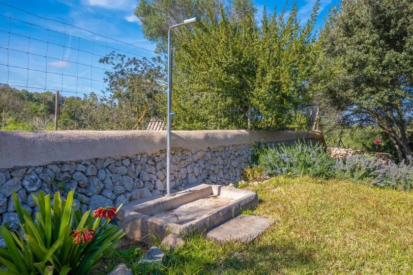 Ducha exterior junto a un muro de piedra, con árboles y vegetación cerca.