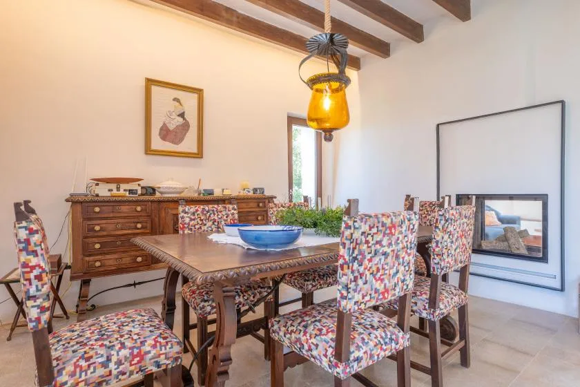 Un comedor con una mesa larga y sillas tapizadas de colores bajo vigas vistas.