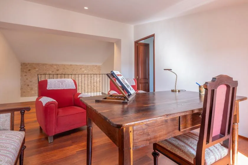 Un escritorio de madera y un sillón rojo amueblan una habitación luminosa con una pared de piedra.