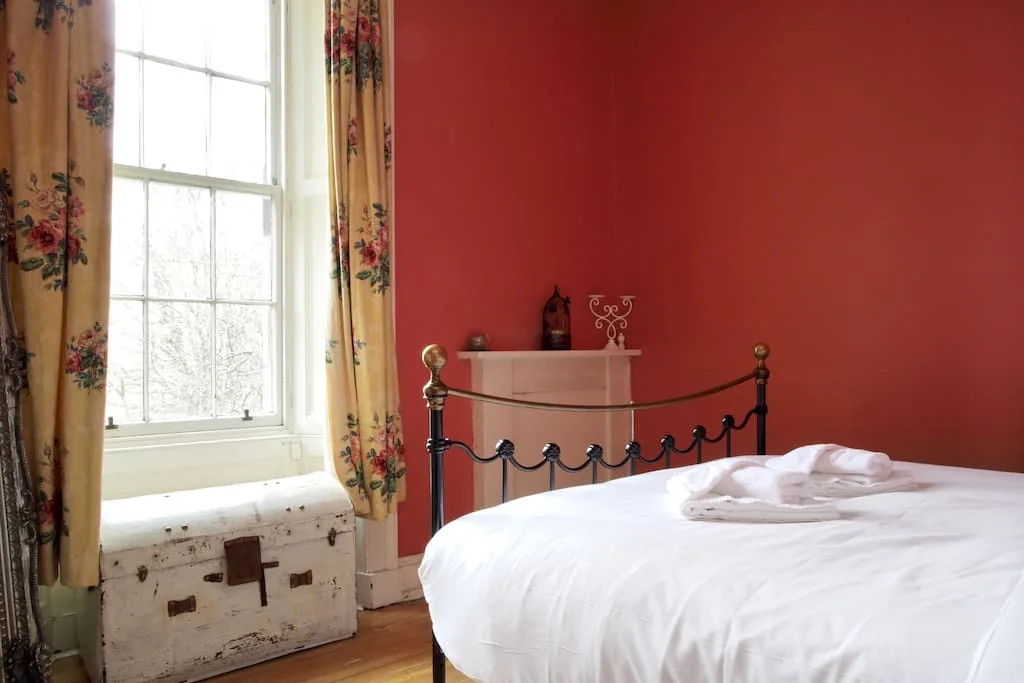 A bedroom with a white bed, a red wall, and floral curtains beside a window.