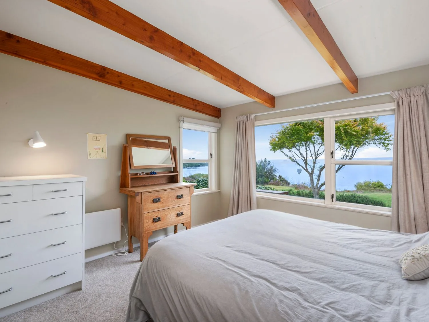 Un dormitorio con una cama, una cómoda blanca, un tocador de madera y ventanas con vistas a árboles y vegetación.