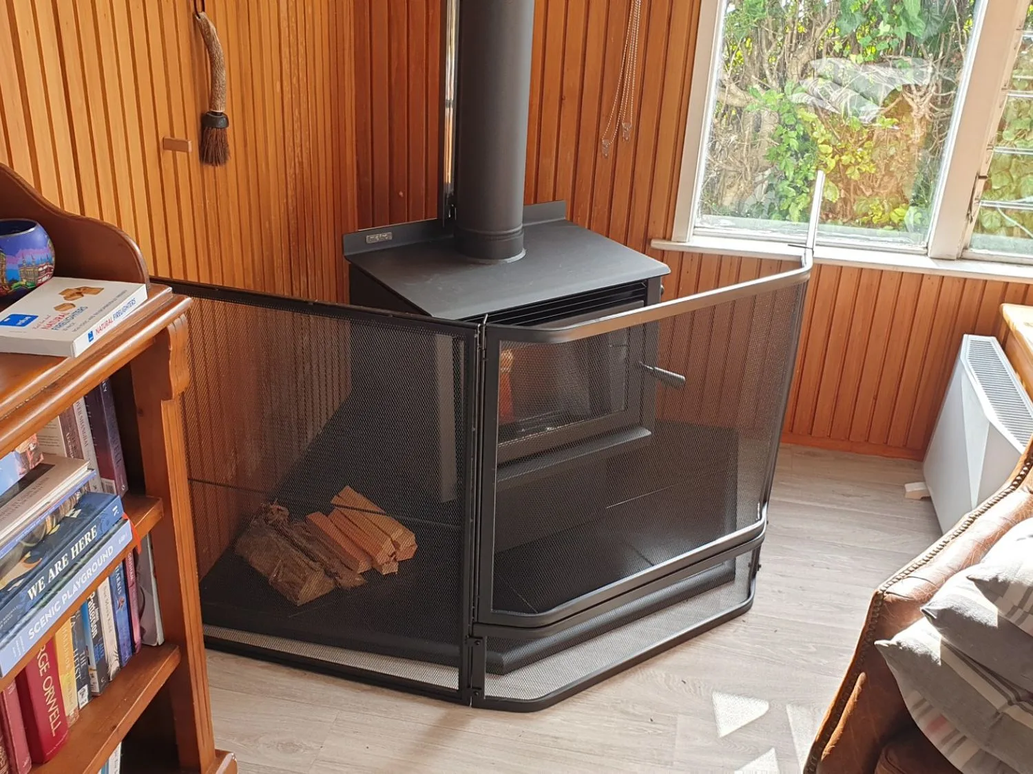 Una estufa de leña con leña y una pantalla de protección en una habitación revestida de madera con una estantería y una ventana en Henley - Omori Holiday Home, en Omori