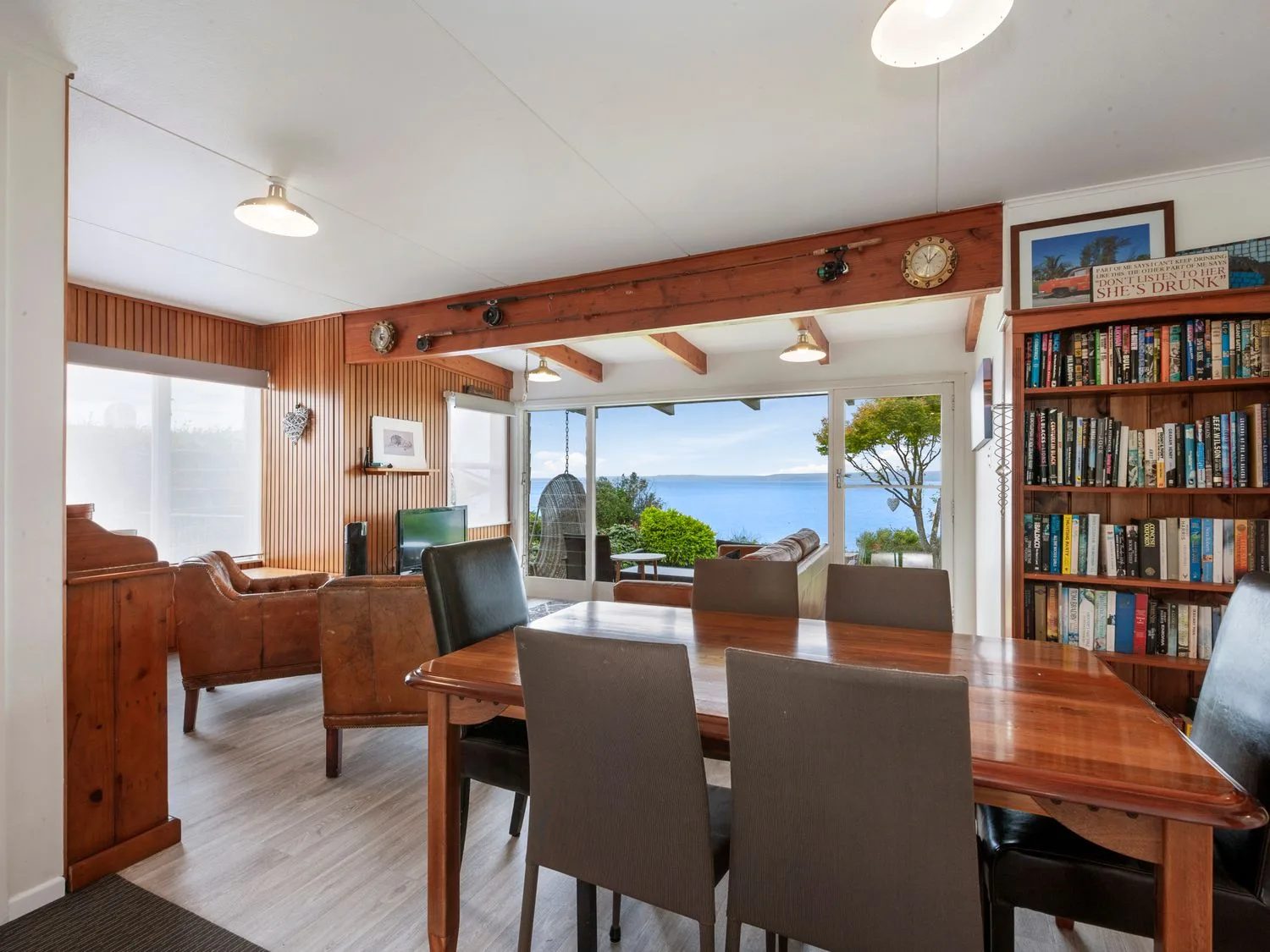 A dining area with a wooden table and six chairs next to a living room with leather chairs and a TV facing large windows showing a water view at Henley - Omori Holiday Home in Omori