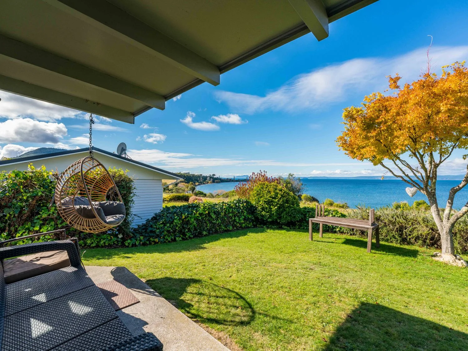 Un patio trasero con una silla colgante, un sofá de mimbre, un banco de madera, un árbol con hojas anaranjadas y vistas al mar en Henley - Omori Holiday Home, en Omori
