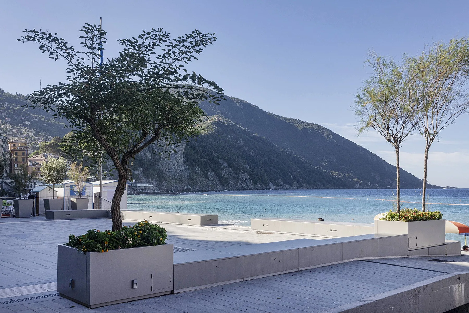 A paved seaside terrace with planters overlooks the beach, blue water, and a mountainous coastline.