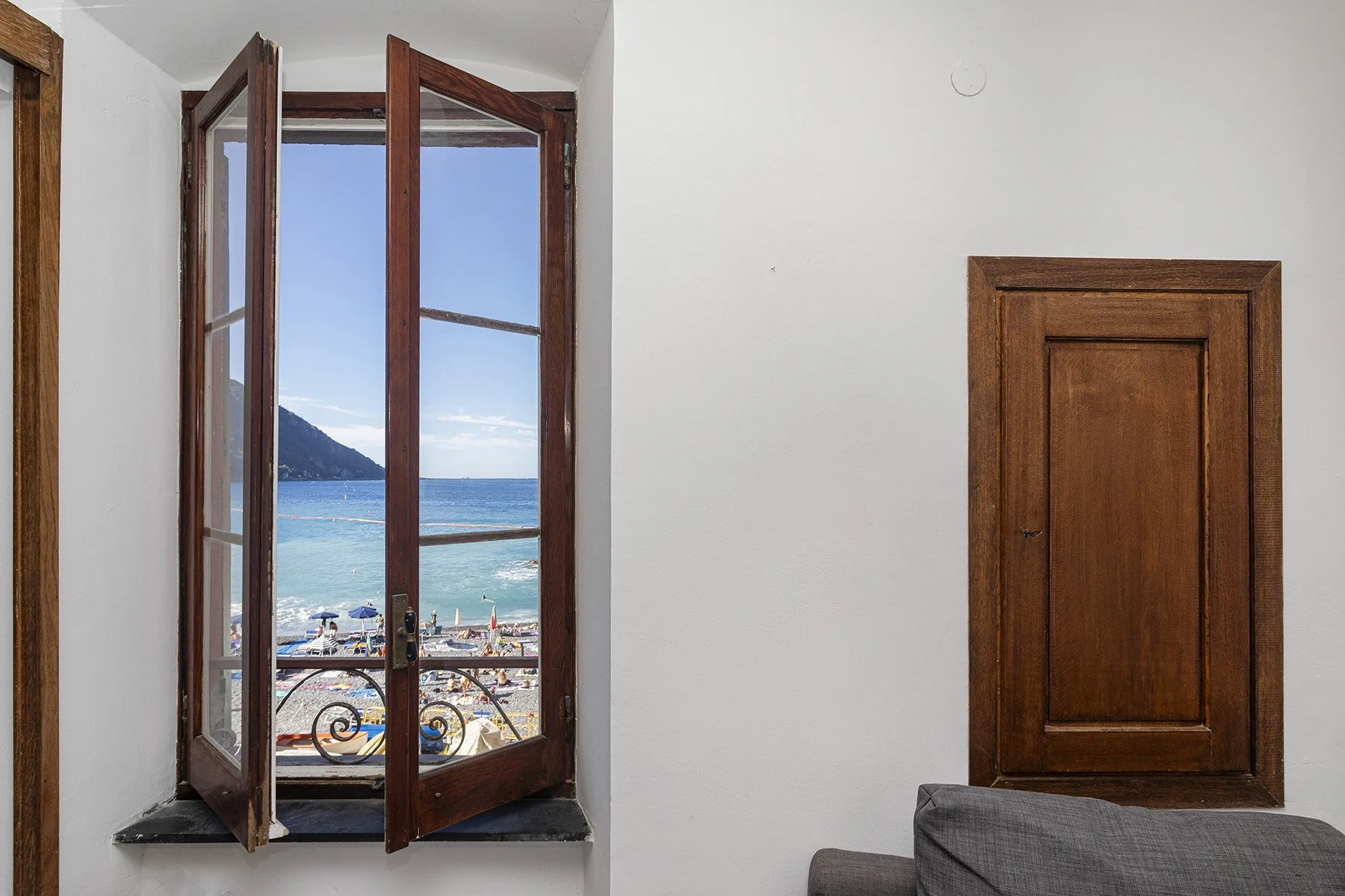 A room with open wooden windows overlooking the sea and a beach.