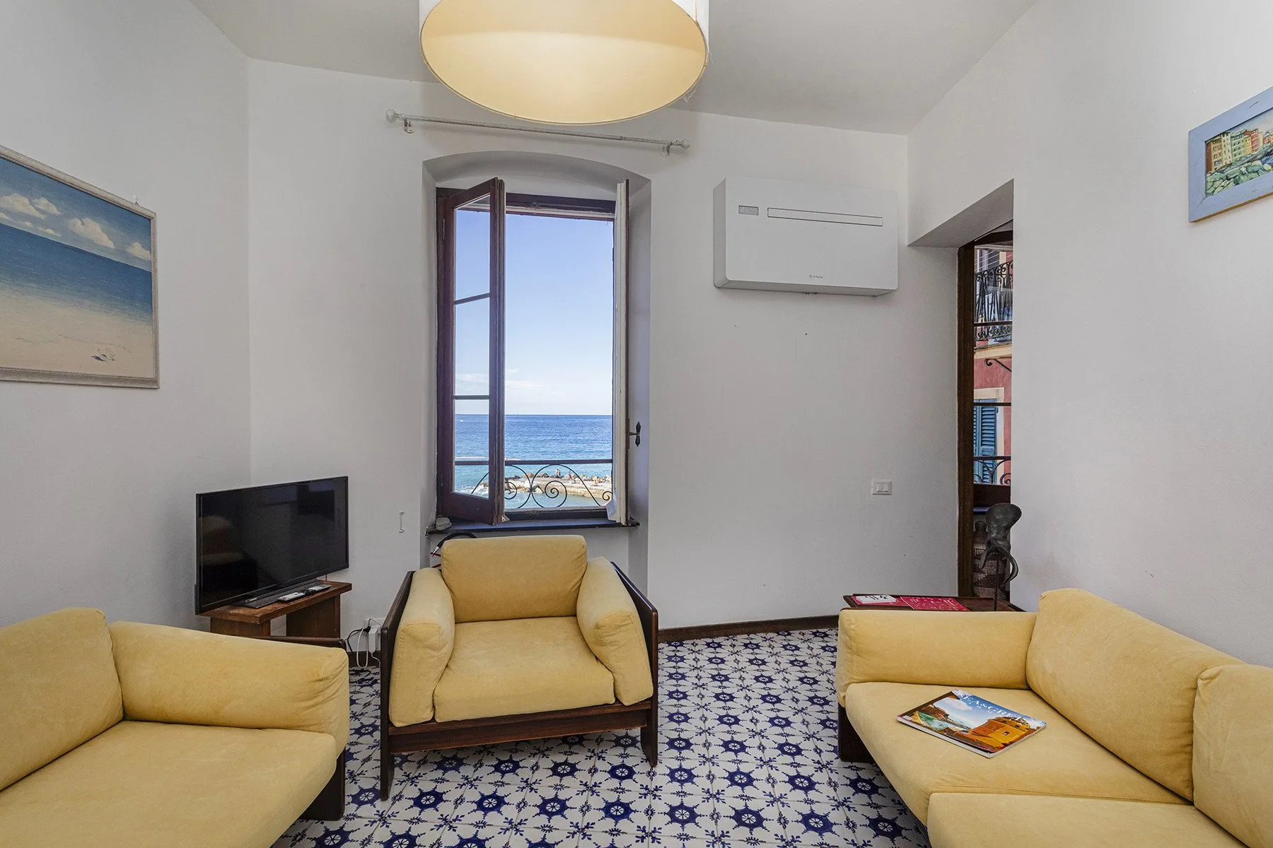 A living room with yellow seating, patterned floor tiles, and a window overlooking the sea.