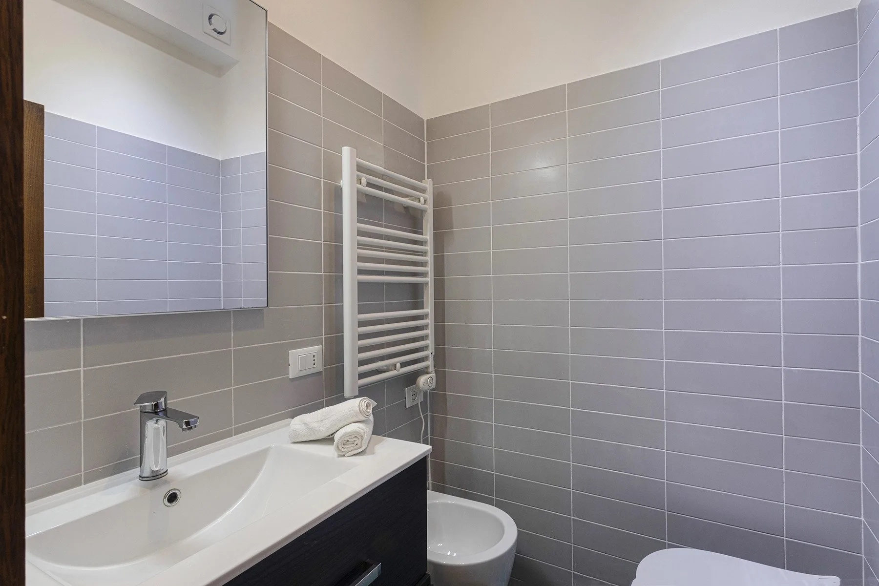 A tiled bathroom with a sink, mirror, and wall-mounted towel warmer.