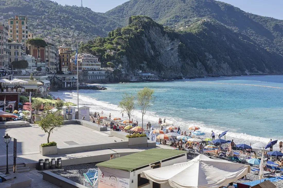 A coastal beach and waterfront promenade sit beneath a steep, green hillside.