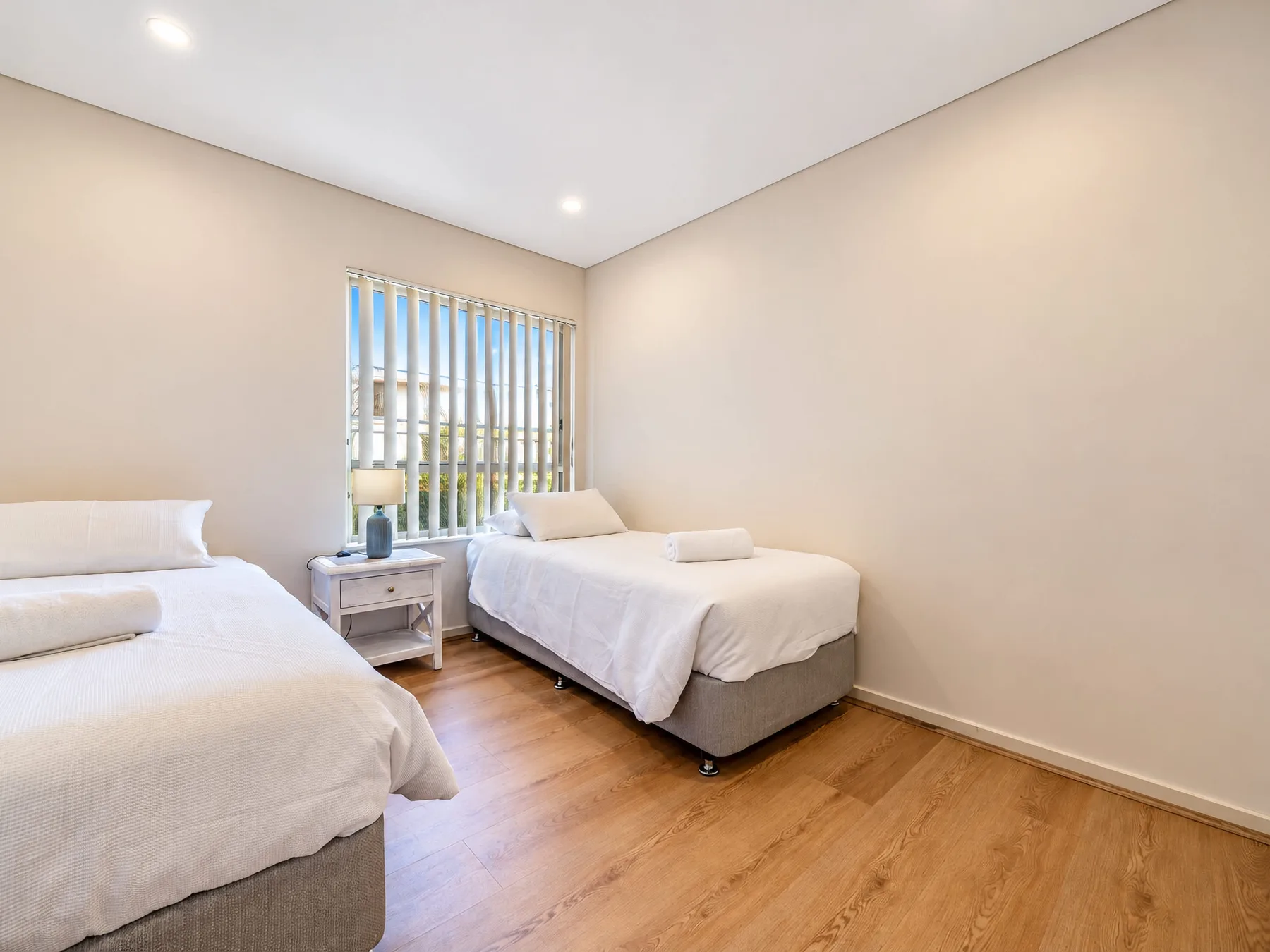 A bright bedroom with two beds and a window with vertical blinds.