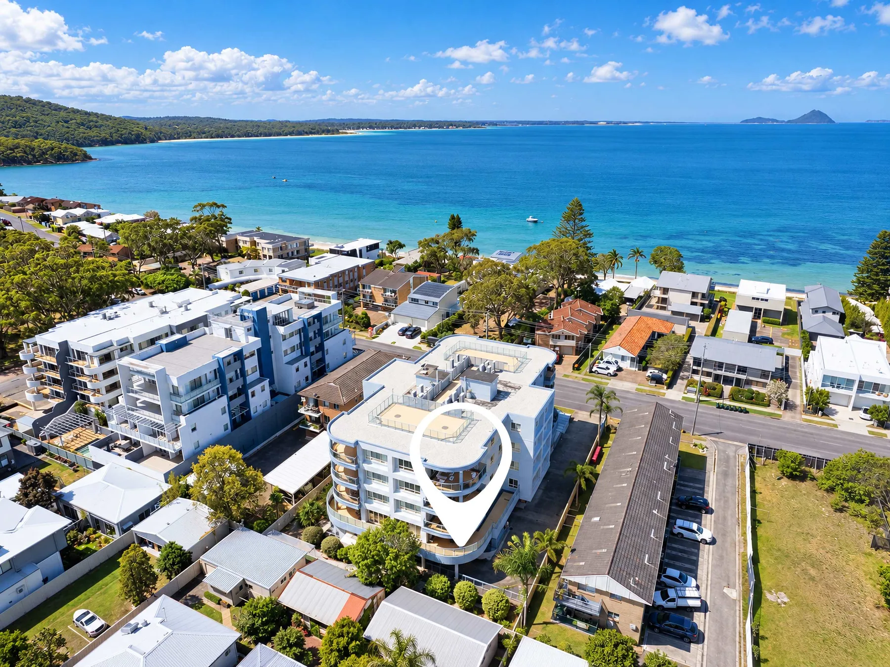 Aerial view of coastal buildings beside a blue bay and tree-lined shoreline.