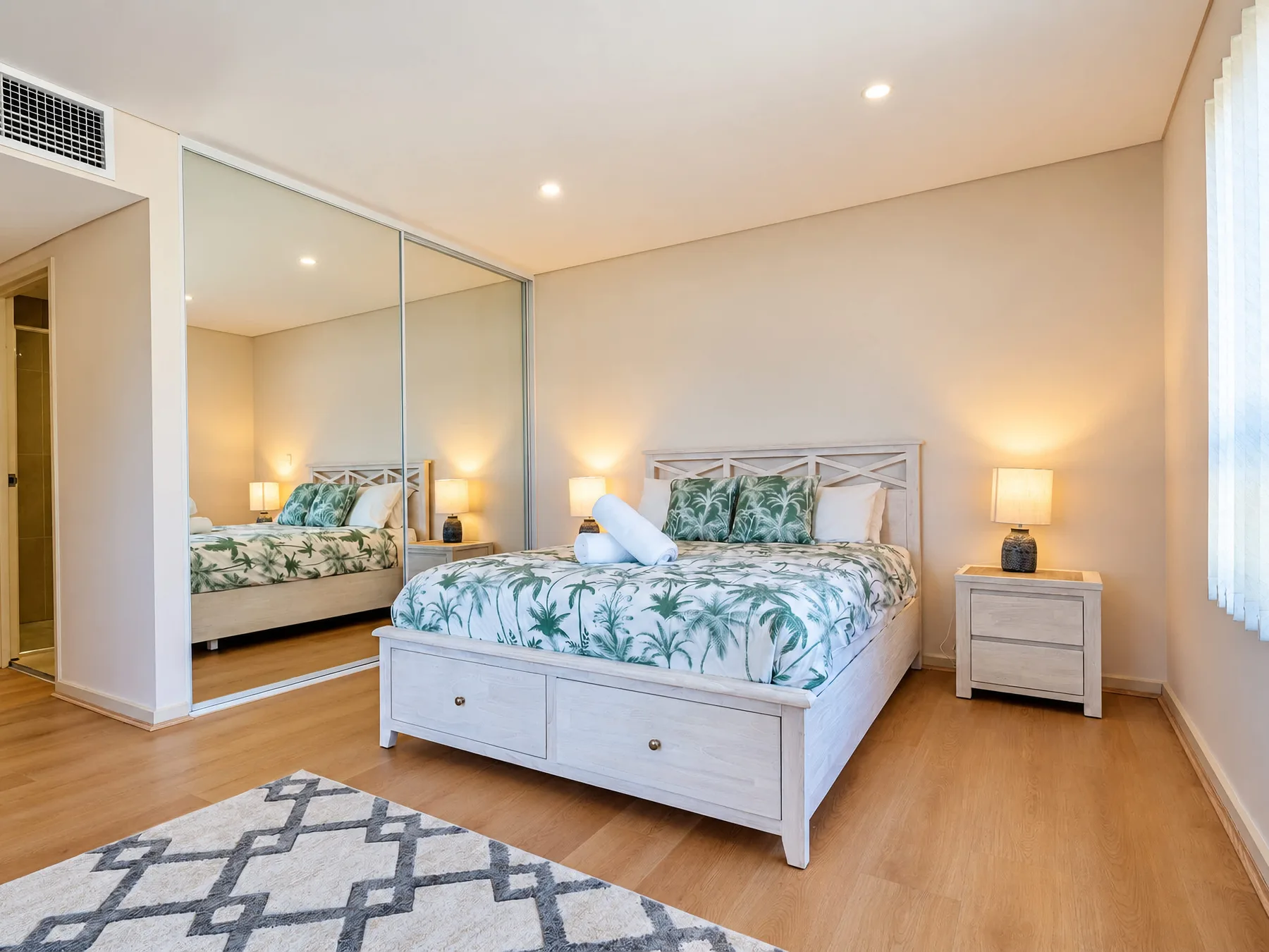 A bedroom with a bed dressed in green palm-patterned bedding and a mirrored wall.