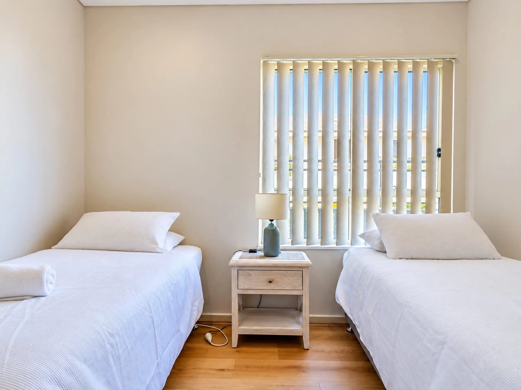 A bedroom with two single beds, a bedside table, and vertical blinds.