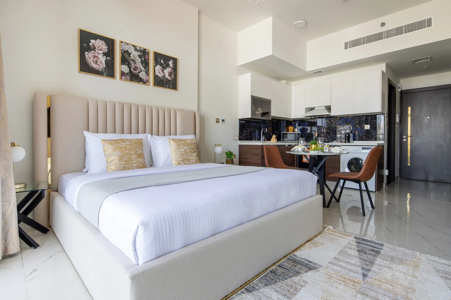 A bedroom with a large bed and a kitchen dining area in the background.