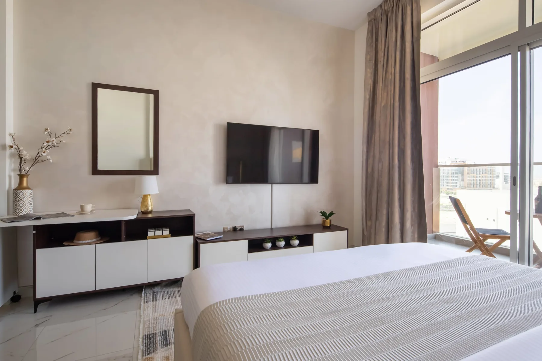 A bright bedroom with a neatly made bed, wall-mounted television, and glass doors to a balcony.