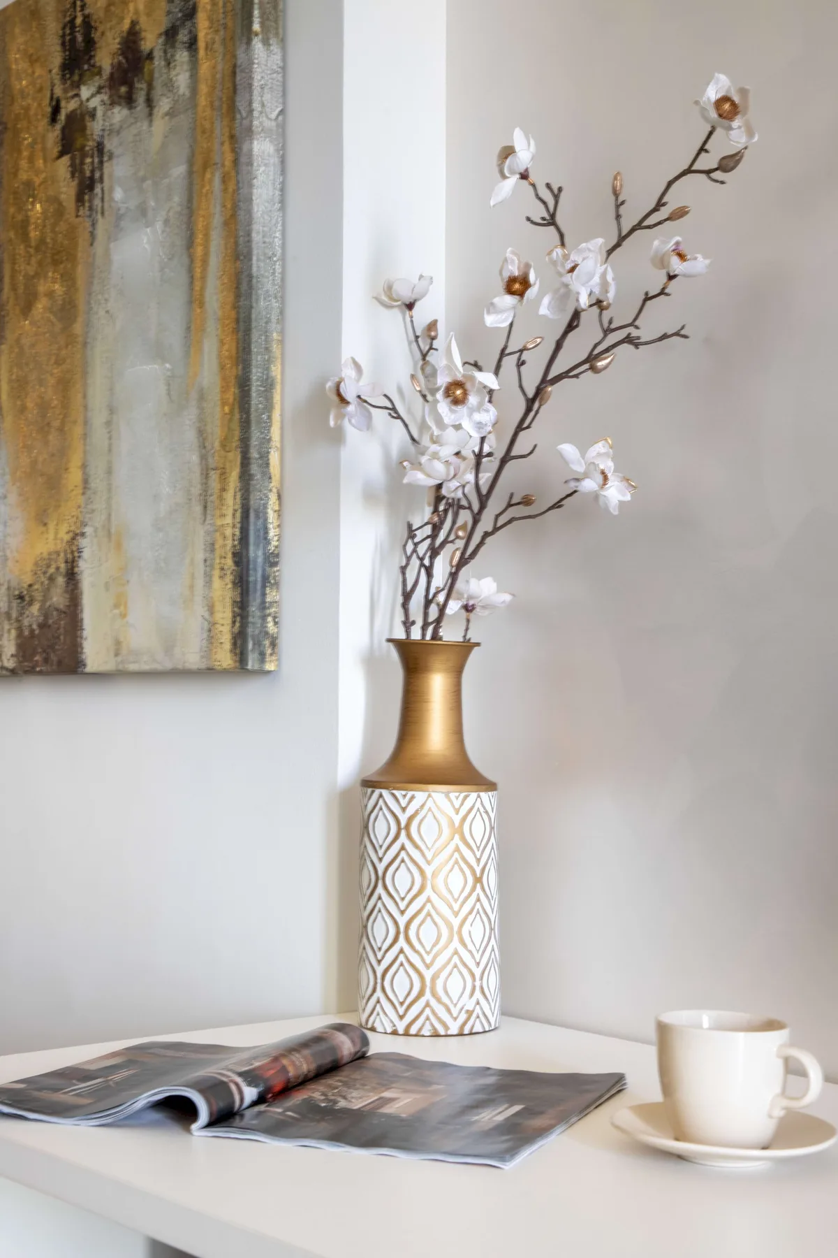 A decorative vase with flowering branches stands beside an open magazine and a cup.