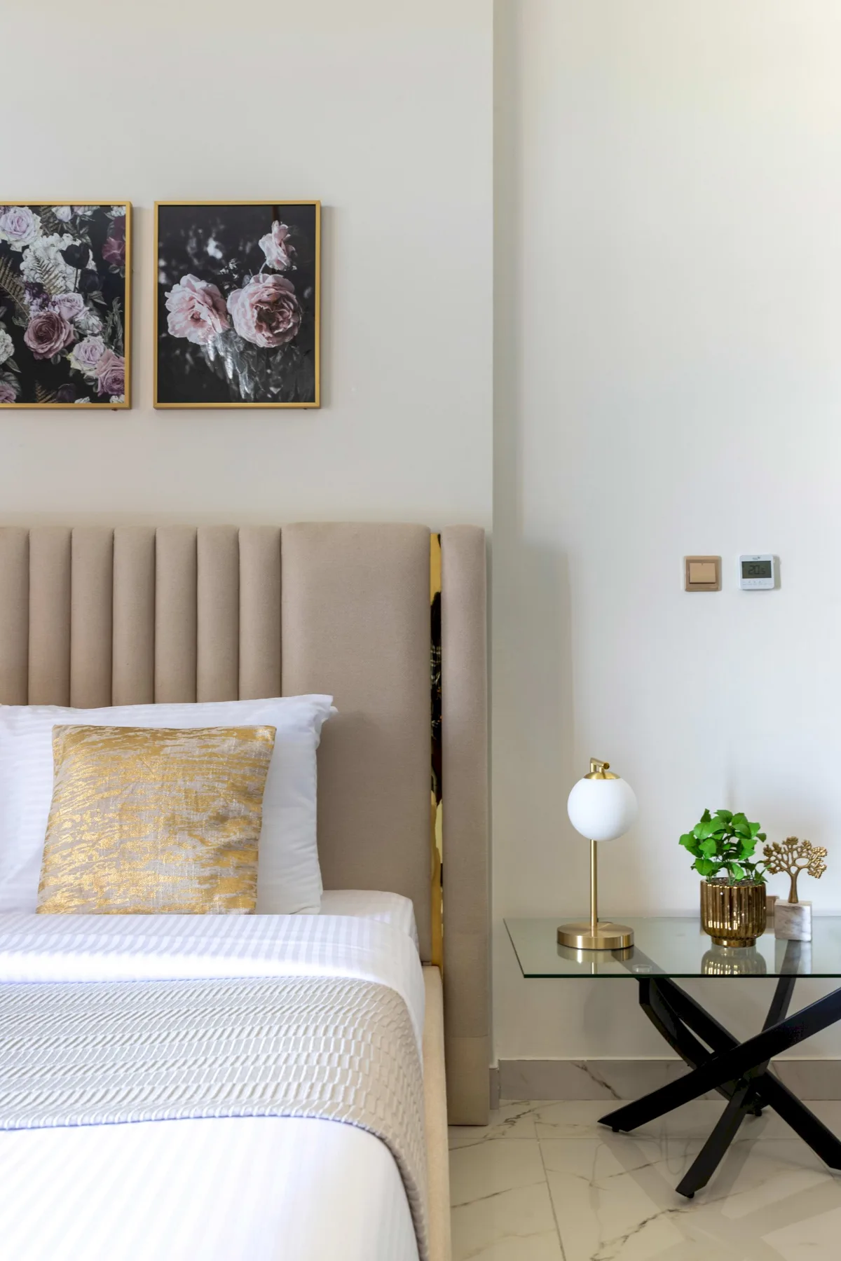 A bedroom with a neatly made bed, floral artwork, and a glass side table.