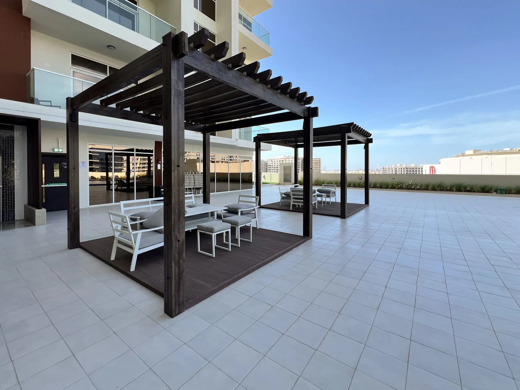 A tiled outdoor terrace features dark pergolas with seating beside a modern building.