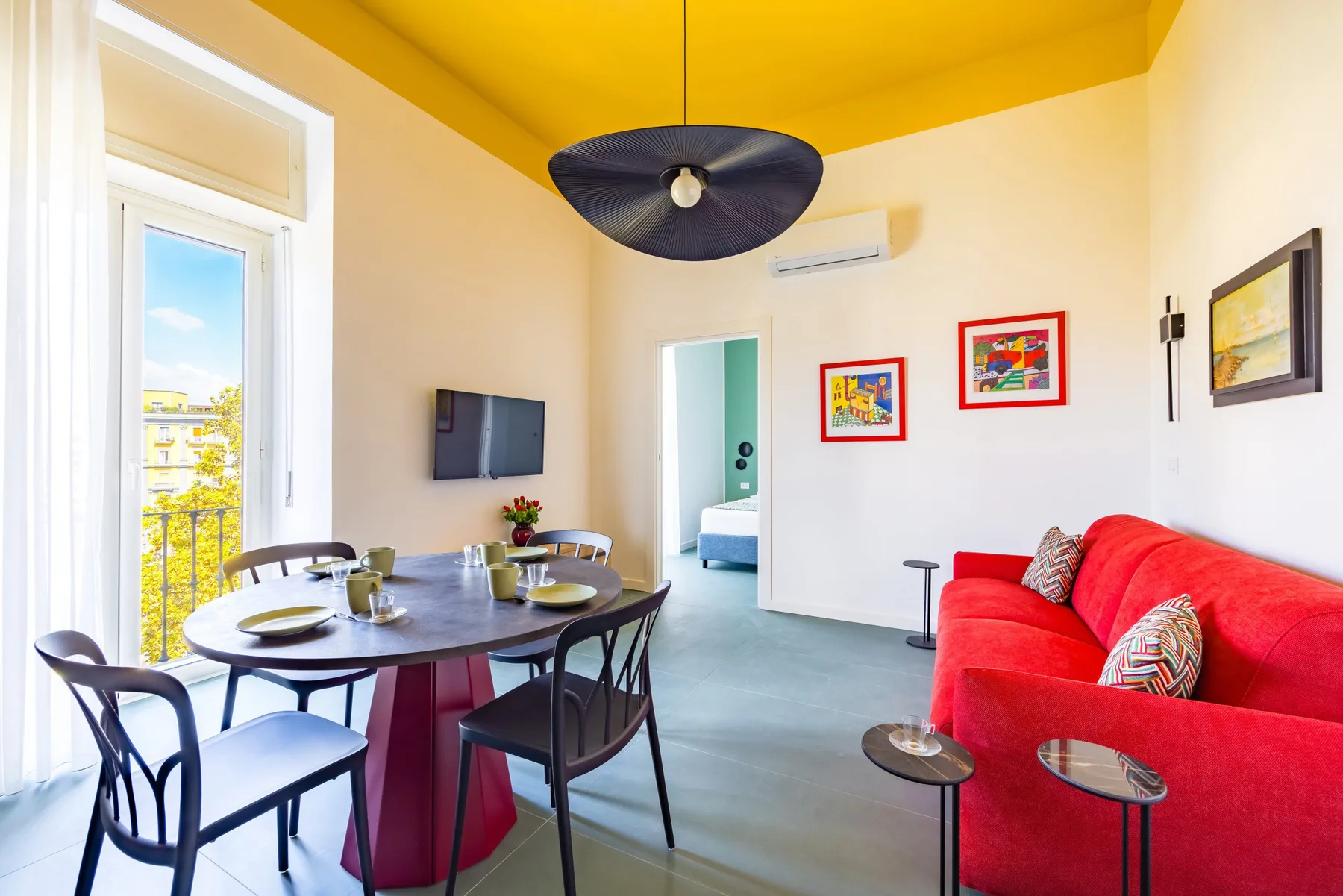 A bright living room with red seating, a dining table, and a balcony door.
