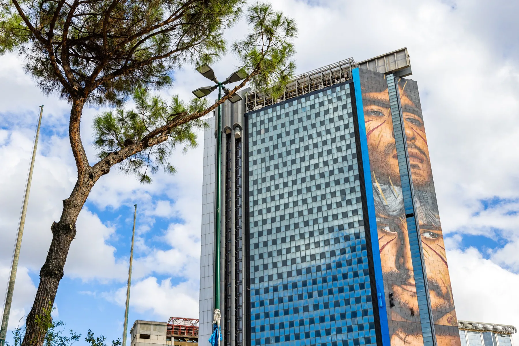 Un edificio moderno y alto con ventanas azules y un gran mural se alza bajo un cielo parcialmente nublado.