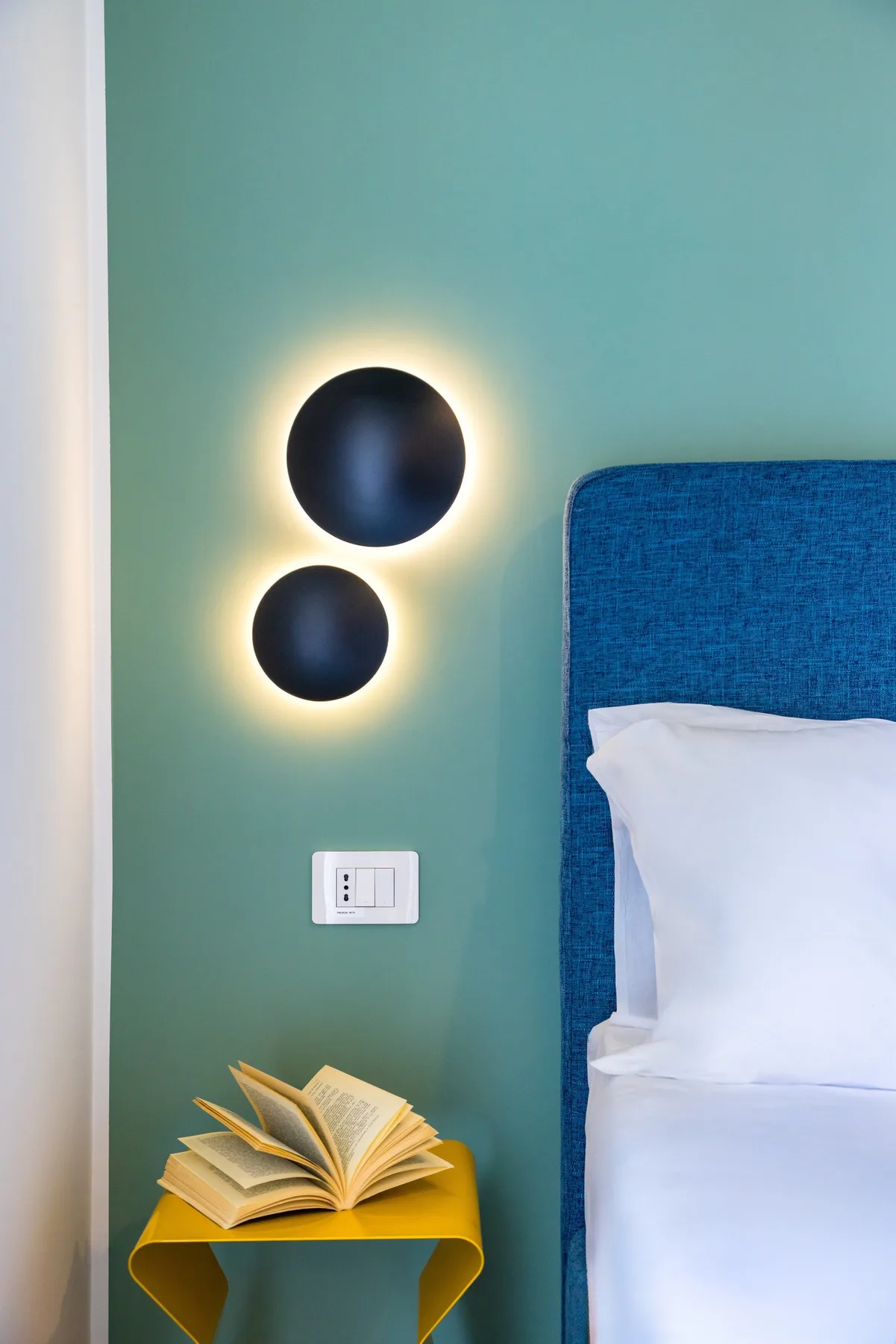 Una cama con cabecero azul está junto a una mesita amarilla y luces de pared.