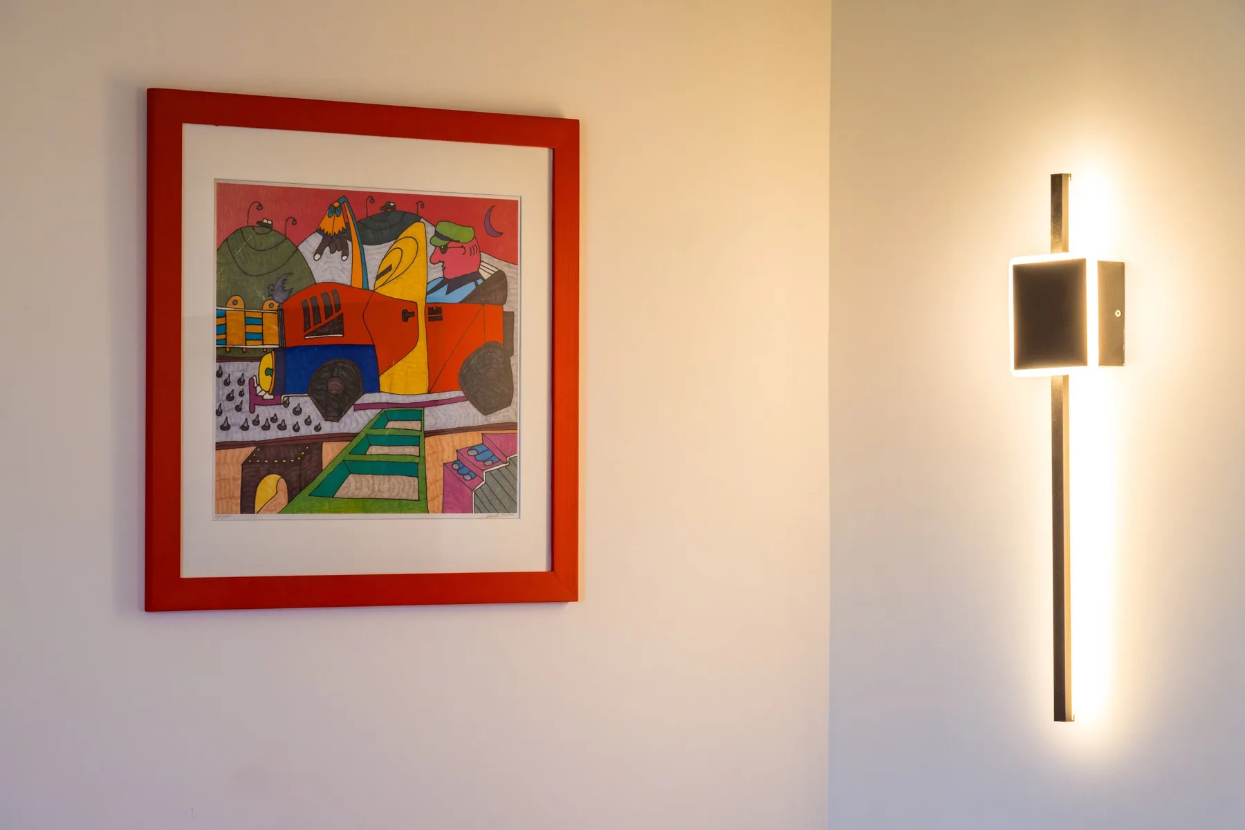 Una obra colorida enmarcada cuelga de una pared clara junto a un aplique.
