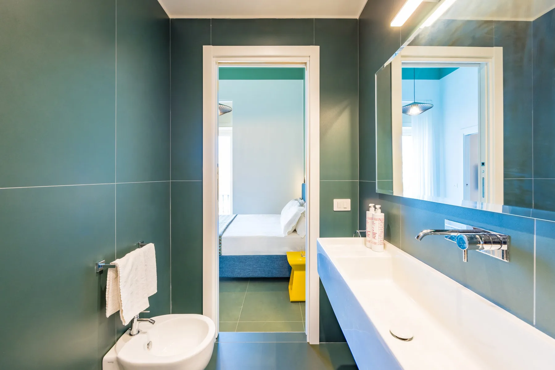 Un baño con un lavabo alargado, espejo y puerta hacia un dormitorio.