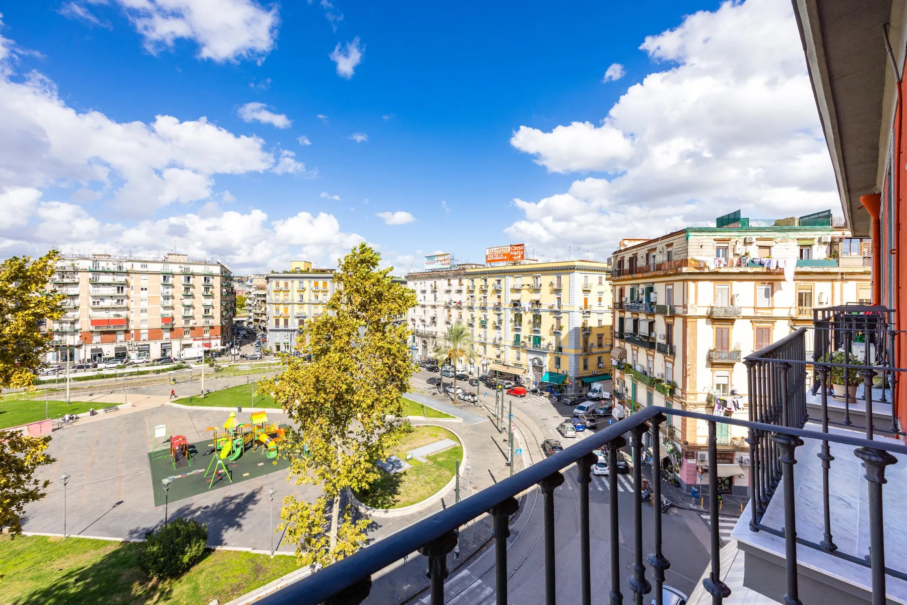 Un balcón da a una soleada plaza urbana rodeada de edificios y árboles.